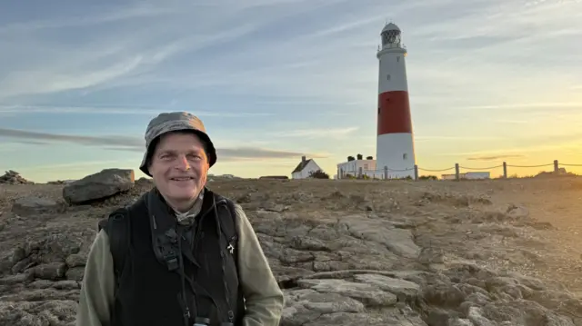 A man wearing a hat and a vest is standing by a lighthouse.