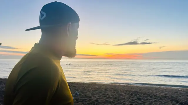 A man wearing a cap is watching the sunset at the beach.