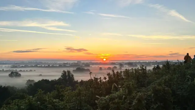 The sun is rising over a misty valley.  There are people watching the sunrise to the right of the image