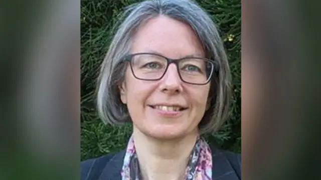 Geraldine Coggins smiles in a headshot picture. She is wearing a floral scarf and a grey jacket in front of a green nature backdrop.