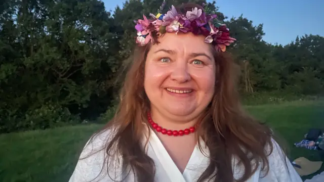A woman wearing a flower crown is sitting on a grassy hill. She is smiling.