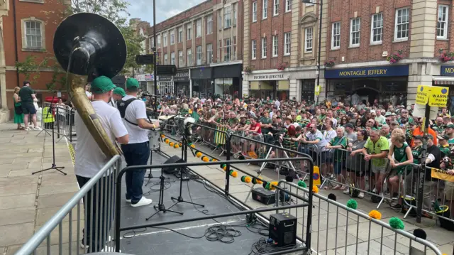 People are standing on a stage playing instruments on the left. To the right is a crowd of people who are watching the performers and who are standing behind a silver barrier.