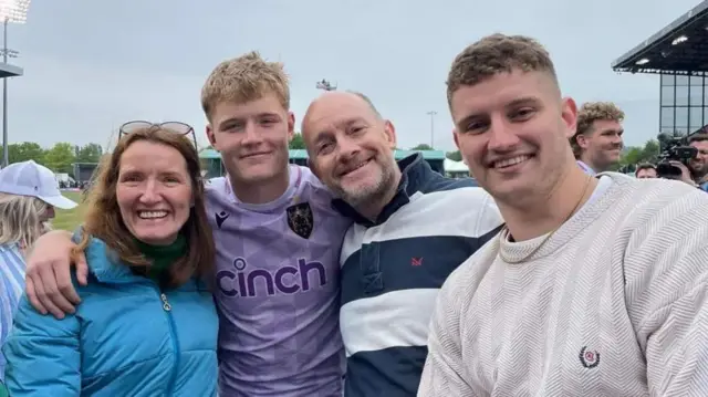 A woman in a blue coat stands smiling next to a man in a purple rugby top. A man in a navy blue and white horizontal polo shirt rests his head on the rugby player's shoulder while a man in a white jumper and gold necklace smiles at the camera. In the background is a rugby pitch and stadium.