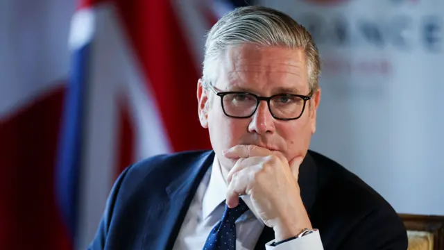 Prime Minister Keir Starmer looking pensive with his head propped up on his left hand. He's wearing a dark suit, white shirt and a blue tie with small white polka dots. In the blurred background, to his right, is a Union Jack