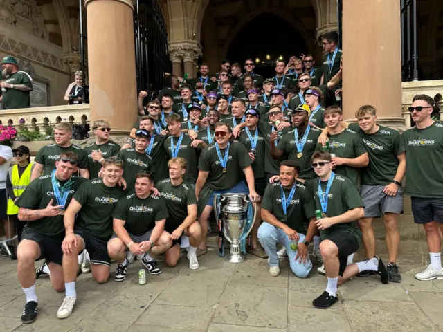 Several Northampton Saints players and club members are standing on a stairway which leads into Northampton's Guildhall. Nearly all of them are wearing medals. They are all standing around a large silver trophy.
