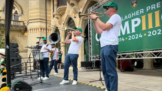 Four men are standing on a stage and are all playing different instruments, including drums and a saxophone. They are all wearing navy trousers and a white T-shirt and are standing in front of a sign which is green.