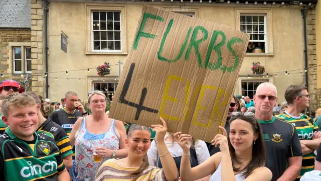 Two women are holding a carboard sign which says FURBS 4EVER in different colours. Several people are standing behind them and are wearing Northampton Saints rugby shirts.