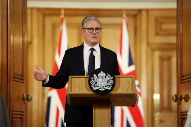 Prime Minister Keir Starmer stands behind a wooden lectern bearing the symbol of the UK. He's wearing a black suit, with a white shirt and black tie, behind him in the blurred background, are two Union flags