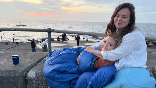 A woman is holding a child who is wrapped up in a sleeping bag. They are sitting by the sea.