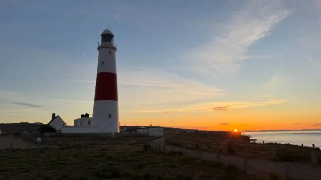 View of a lighthouse by the sea with the sun rising on the horizon.