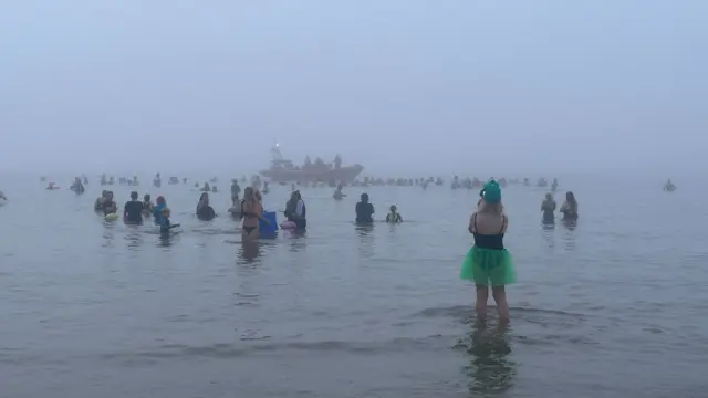 People are standing in the sea.  It is very foggy. There is a boat in the background