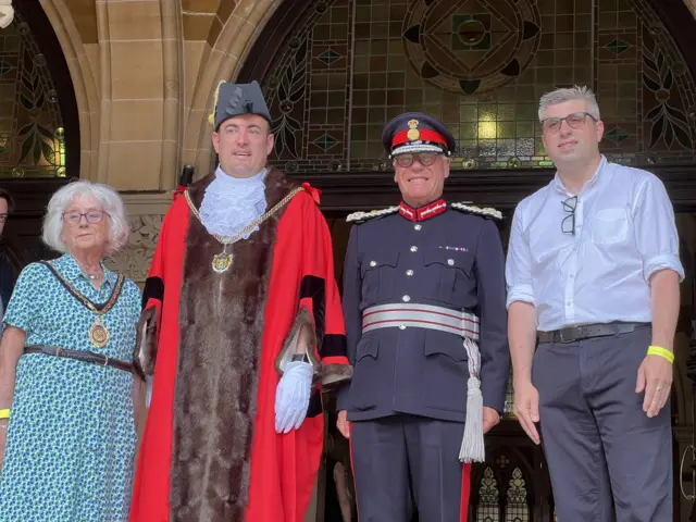 Four people are standing next to each other in a doorway, including the towns the mayor and a councillor. They are all smiling and looking towards a crowd.