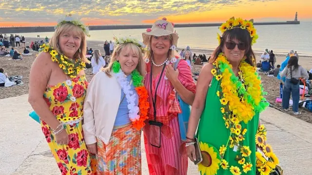 Four women in colourful clothes are by the sea.