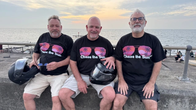Three men are sitting on a ledge by the beach. They are wearing black t-shirts that say "Chase the Sun"