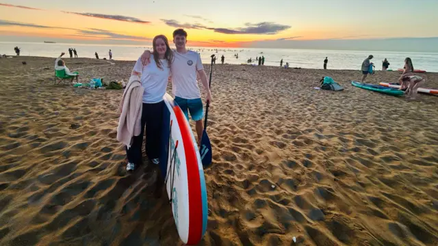 A boy and a girl have their arms around each other next to a paddleboard on a beach at sunrise.