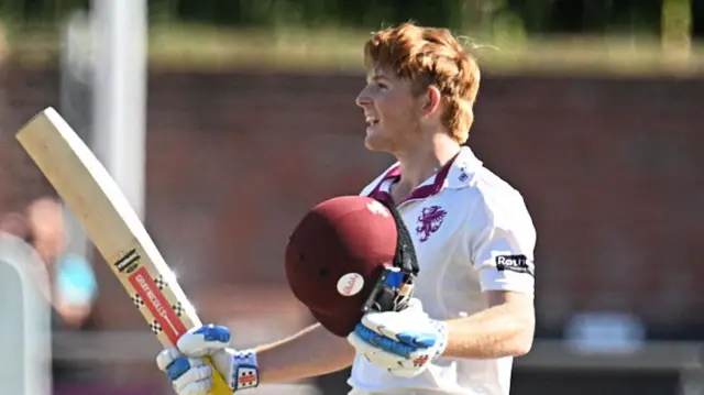 Thomas Rew holds his helmet in his hand and waves his bat in the air to celebrate his century