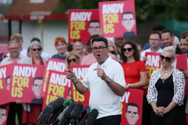 Andy Burnham in a white polo and black jeans addresses supporters at a victory rally in Ashton-in-Makerfield. Behind him, to the left, is his wife in a white shirt with black polka dots over a black dress. Next to her are several supporters holding red Andy for Us signs