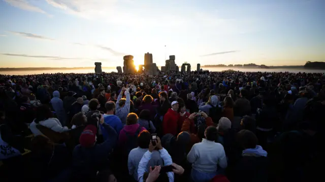 A crowd has gathered in front of Stonehenge. Some are taking pictures or posing for photos.