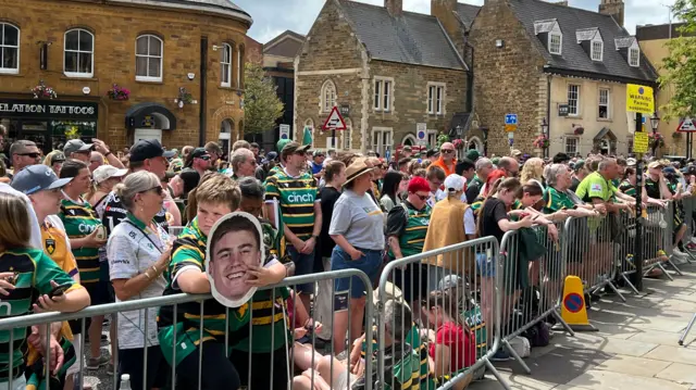 Hundreds of people are gathering in a town centre and are standing behind a metal fence. Most of them are wearing Northampton Saints rugby shirts.