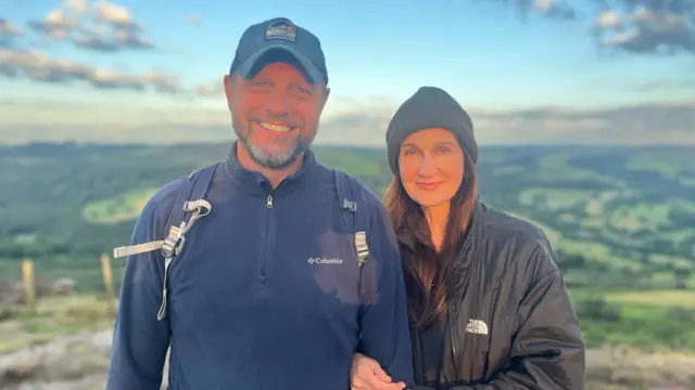A couple is standing on top of a hill, smiling at the camera.