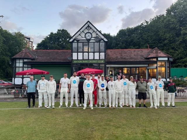 A group outside a cricket club. Most are dressed in cricket gear and are smiling towards the camera.