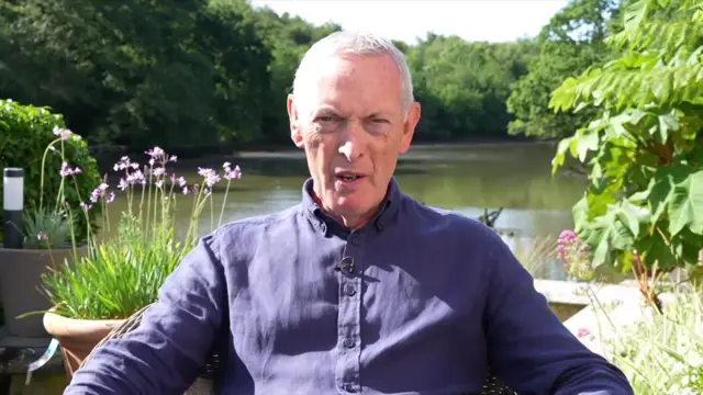 Former defence secretary Lord Hutton in a blue linen shirt sitting on a deck overlooking a river, shrubbery and flower pots around him