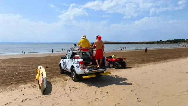 Two lifeguards stand on the back of an RNLI pick up truck on a sandy beach overlooking the sea on Barry island. A surf broad is propped up to the left of the car and an ATV is seen to the right