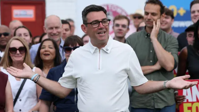 Burnham, wearing a white polo shirt, opens his arms with a crowd behind him
