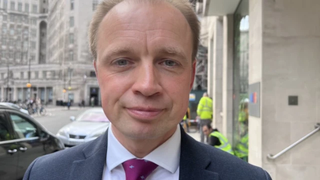 A man stood close to the camera in a city centre looking into the camera. He is wearing a suit.