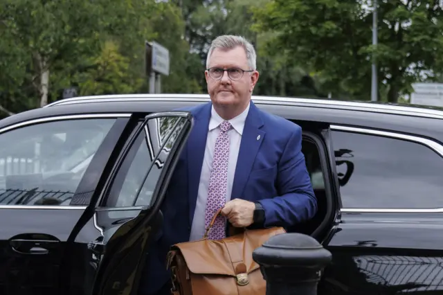 Donaldson is getting out of a black car. He is carrying a brown brief case. He is wearing a navy suit, white shirt and pink tie.
