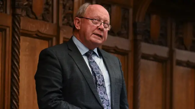 An older man with grey hair at the side of his head and bald on top is standing in front of a wooden panelled wall. He is wearing a dark suit, dark patterned tie and a grey shirt.