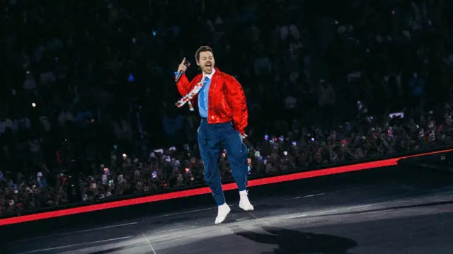Harry Styles wears a red bomber jacket, light blue shirt, floral tie and navy trousers while performing on stage. He can be seen holding his mouth open with a microphone in front of a crowd of people holding their phones