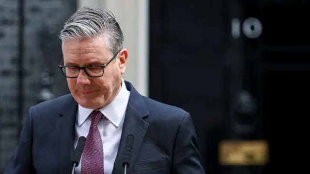Keir Starmer is pictured speaking behind a lectern at Downing Street, wearing a suit and tie. His head his bowed as he looks down.