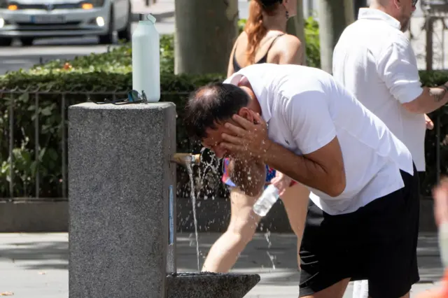 A middle aged man with black hair wearing a white shirt and black shorts washes his face in a public water fountain in the middle of the street
