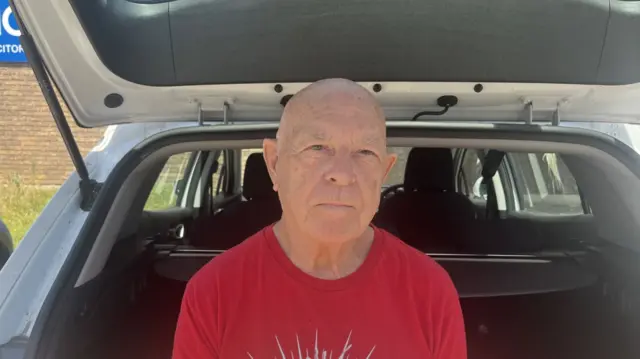 Dave Holmes wears a red t-shirt and sits against the boot of a car