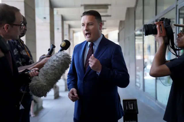 Wes Streeting, wearing a navy suit, gestures as he speaks to a number of journalists holding microphones outside Portcullis House in Westminster