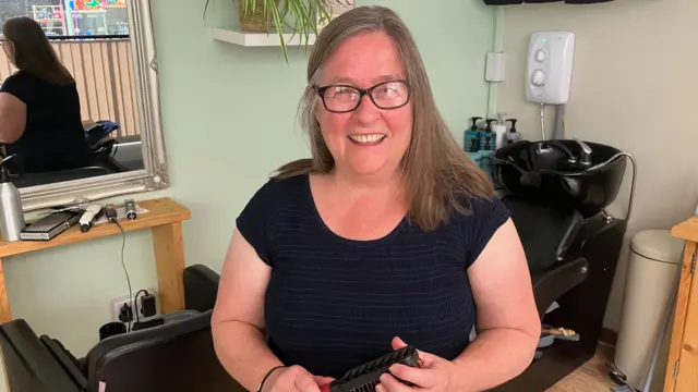 Jo has long brown hair and is holding a hairbrush. She is stood in her hairdressers. A chair with a sink can be seen behind her on the right and a mirror can be seen on the left.