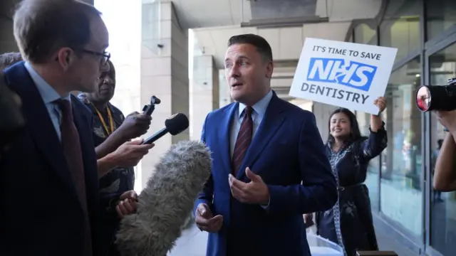 Wes Streeting speaks to the BBC's Chris Mason outside Portcullis House in Westminster. A protester holds a sign saying 'it's time to save NHS dentistry' behind Streeting