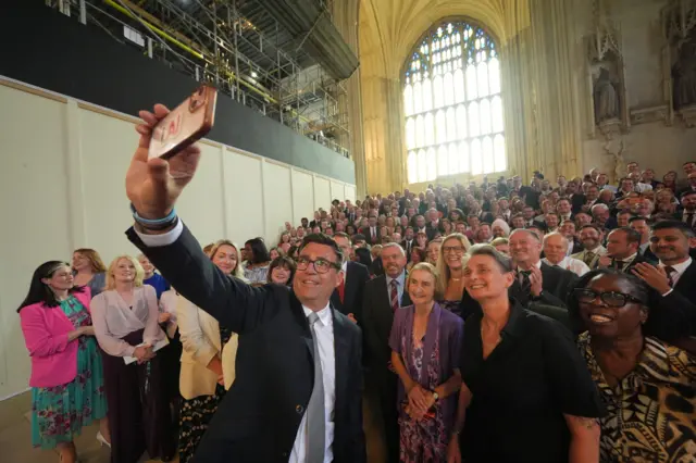 Andy Burnham is holding an iPhone, taking a selfie with a large group of smiling Labour MPs in Westminster hall