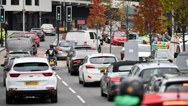 Car traffic in the centre of Bristol. Cars and a motorcyclist are waiting at a set of traffic lights.