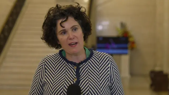 Midshot ofClaire Hanna, wearing blue patterned jacket. Has short black hair.