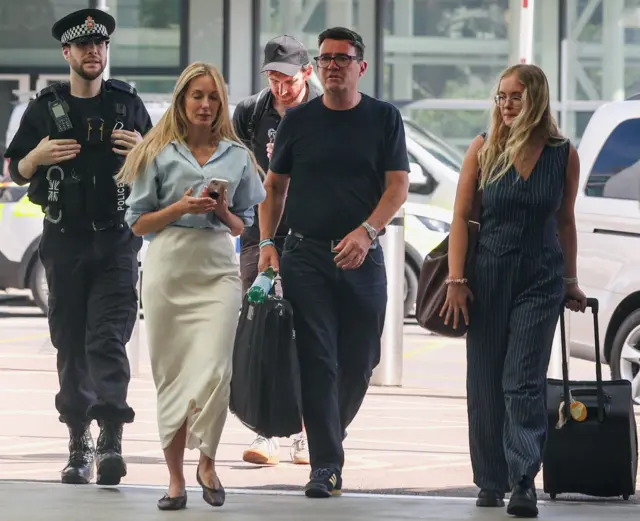 Flanked by two police officers and two female colleagues, Andy Burnham, wearing a dark top, jeans and Adidas trainers, arrives at a sunny Manchester Piccadilly before his train to London Euston
