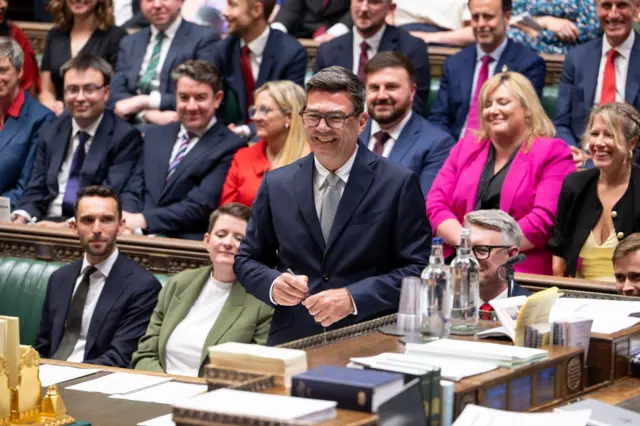 Andy Burnham, wearing a suit and tie, laughs as he is sworn in as the MP for Makerfield in the House of Commons. Behind him are more than a dozen smiling MPs