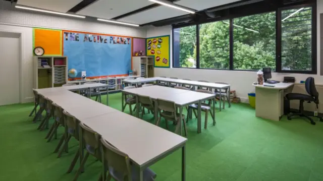 An empty classroom with green carpet and grey desks. There are trees outside the large window, and a blue display board which says 'The Active Planet'