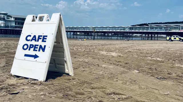 A 'cafe open' a frame sign stands on Weston beach with the sea and the pier in the background.