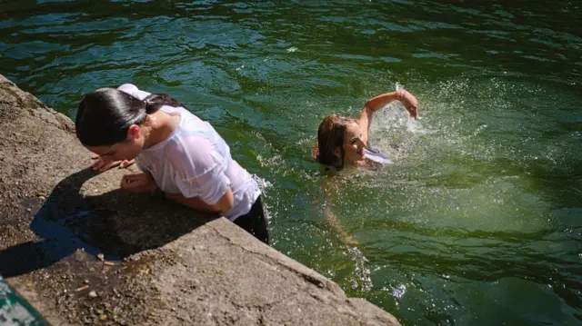 Teenage girls swimming in green water during a heatwave. One of them is wearing a white t-shirt and pulling herself up out of the water.