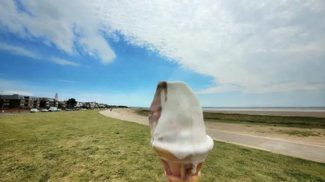 A melted ice cream and chocolate flake on a waffle cone is being held by someone off camera in the middle of a grassy field next to the beach
