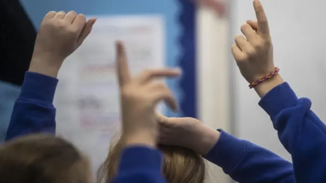Unidentified children in a classroom raising their hands.