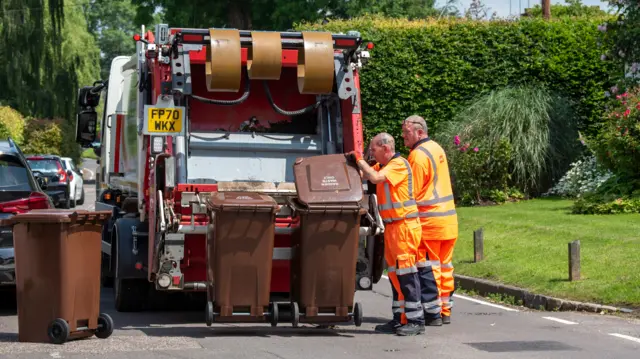 Two bin men wearing orange hi-vis overalls, emptying a brown bin of garden waste into the back of a bin lorry. It is a warm sunny day.