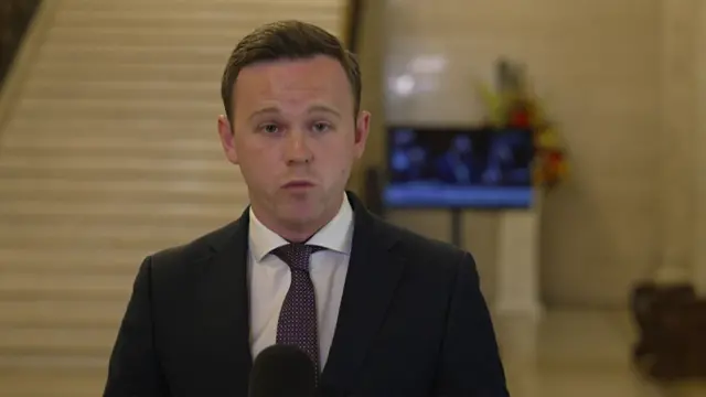 A man wears a navy suit jacket with a white shirt and purple tie. He has short brown hair.